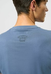 Mėlynas medvilninis marškinėliai su apvaliu iškirpimu ir tekstūruotu logotipu ant nugaros, kuriame rašoma "AUTENTIŠKAS nuo 1890 m. USPA."