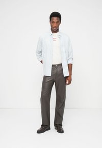 Νεαρός άνδρας που στέκεται φορώντας ένα ανοιχτόχρωμο ριγέ πουκάμισο πάνω από λευκό T-shirt, σκούρα καφέ δερμάτινο παντελόνι και σκούρα καφέ παπούτσια σε λευκό φόντο.