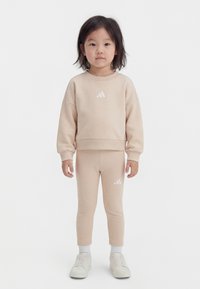 Mlad otrok stoji, oblečen v bež Adidas trenirko in nogavice ter bele superge na belem ozadju.