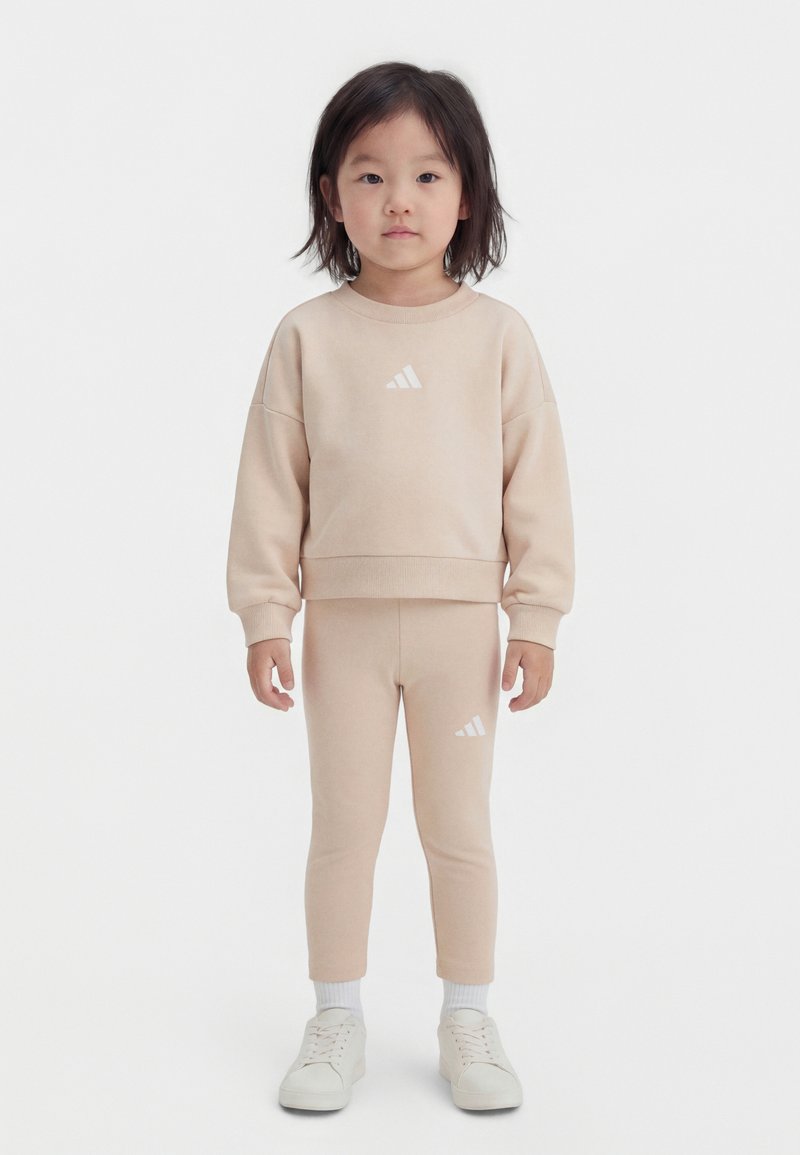 Mlad otrok stoji, oblečen v bež Adidas trenirko in nogavice ter bele superge na belem ozadju.