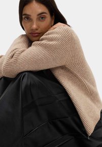 Světle béžový hrubě pletený svetr s žebrovanou texturou, přeložený přes černou plisovanou sukni. Styly zdůrazňují pohodlí a vrstvený design.