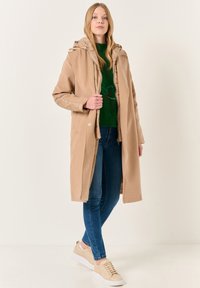 Cappotto lungo beige con cappuccio, chiusura a zip e bottoni, indossato sopra un maglione verde con ricami floreali, abbinato a jeans blu e sneakers.