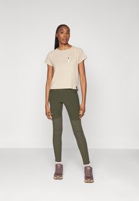 Beige short-sleeve top with a chest pocket, paired with dark green leggings featuring knee accents. Model wears brown outdoor shoes.