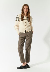 Cardigan bianco in maglia con ricami floreali sulle maniche, bottoni in legno, abbinato a pantaloni a scacchi marroni e mocassini neri.