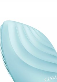Silicone ansiktsrengöringsborste i ljusblått, med vågiga fåror på ena sidan och mjuka borst på den andra. Inkluderar varumärkesgravyr.