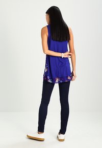 Fioletowy top bez rękawów z kwiatowym brzegiem, noszony na ciemnoniebieskich obcisłych dżinsach i białych tenisówkach, charakteryzujący się prostym krojem i casualowym stylem.