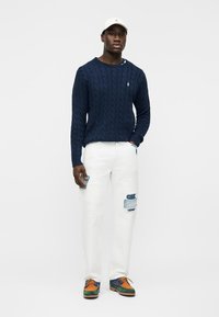 Homme portant un pull en maille torsadée bleu marine, un jean blanc déchiré avec des patches, une casquette blanche et des chaussures multicolores vertes, orange et bleues, debout.