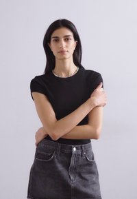 Črna majica s kratkimi rokavi in okroglim izrezom, v kombinaciji s temnimi jeans krilom. Model stoji s prekrižanimi rokami, proti enotni ozadju.