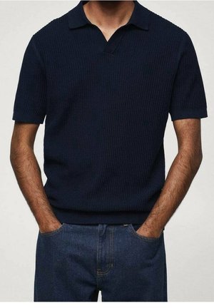 Mand iført en mørkeblå kortærmet strikket polotrøje med krave og blå jeans, hænder i lommer, foran en ensfarvet lys baggrund.