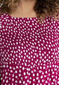 Mujer vestida con un vestido magenta con pequeños patrones florales blancos y azul claro, y con tejido recogido en el pecho y los hombros.