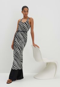 Schwarz-weiß gestreift, ärmelloses Maxikleid mit figurnahem Taillenbereich und schwingendem Saum, kombiniert mit schwarzen flachen Sandalen vor einem minimalistischen Hintergrund.