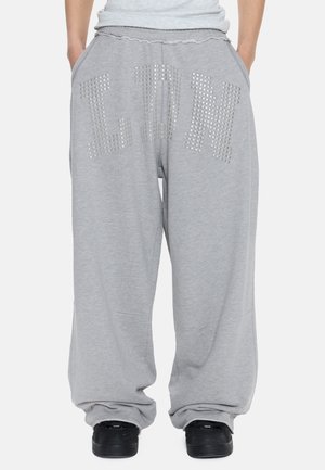 Άτομο που φοράει χαλαρό γκρι παντελόνι φόρμας με καρφωμένα γράμματα "LDN" μπροστά, συνδυασμένο με μαύρα αθλητικά παπούτσια και τα χέρια στις τσέπες.