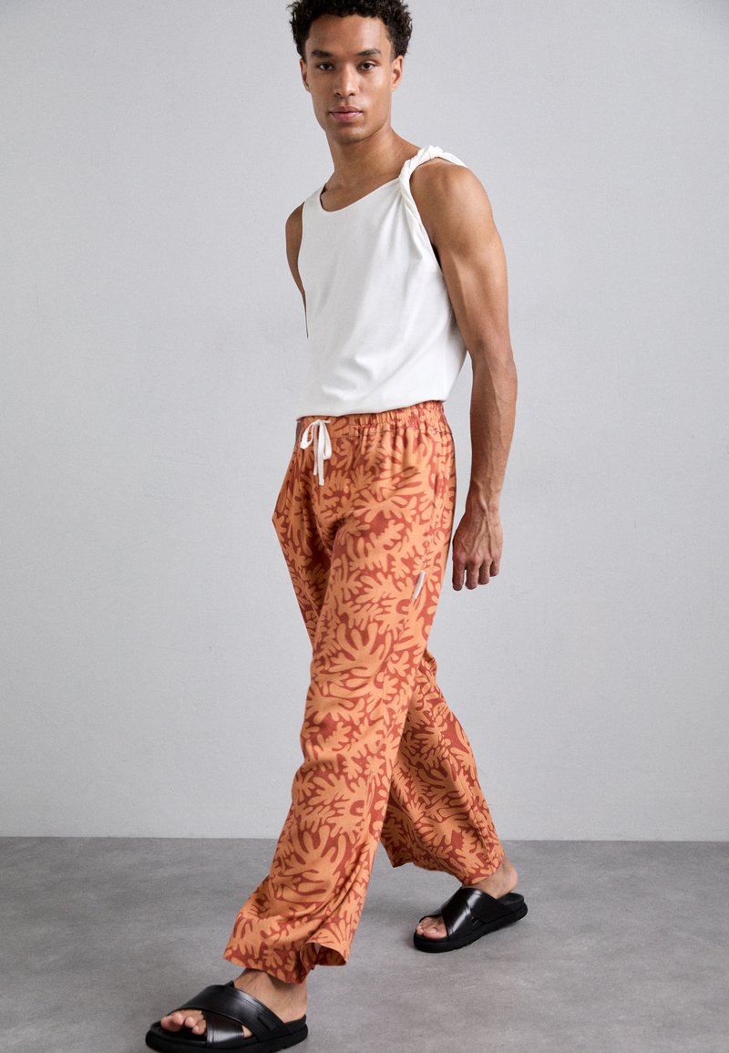 White sleeveless tank top paired with orange patterned pants featuring a drawstring waist. Black slide sandals complete the look.