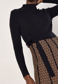 Haut noir côtelé à manches longues rentré dans une jupe à motifs taille haute avec ceinture noire nouée, porté par une personne sur un fond neutre.