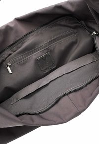 Interno di una borsa in tessuto nero con tasca con cerniera e sezione piatta. Texture liscia con un leggero luccichio. Patch del logo visibile sul rivestimento.