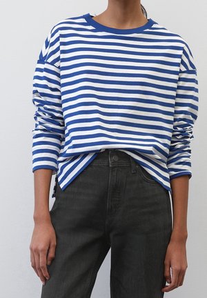Personne portant une chemise à manches longues rayée bleu et blanc horizontalement, rentrée dans un jean noir taille haute, se tenant devant un fond uni.