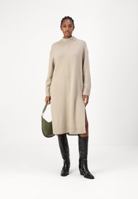 Neulottu beige mekko, jossa on korkea kaulus, resorikätköt ja helma. Yhdistetty kiiltävien polven yli ulottuvien mustien saappaiden ja vihreän olkalaukun kanssa.