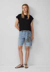 Schwarzes, kurzes, lockeres Hemd, kombiniert mit hellblauen Denim-Shorts mit umgeschlagenem Saum und einem gemusterten Schal, der um die Taille gebunden ist. Schwarze Sandalen.