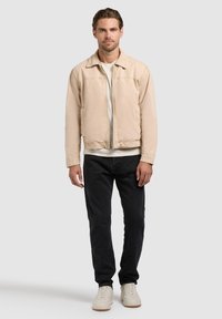 Homme debout face à l'avant portant une veste beige, un t-shirt blanc, un pantalon noir et des baskets blanches sur un fond gris uni.