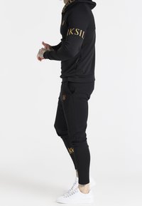 Chándal atlético negro con acentos en logo dorado. Presenta una sudadera ajustada y pantalones ceñidos, hechos de un tejido suave, combinados con zapatillas blancas.