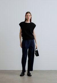 Zwart gebreid topje met korte mouwen, gecombineerd met marineblauwe broek. Model draagt een kleine zwarte handtas en zwarte schoenen. Effen achtergrond.