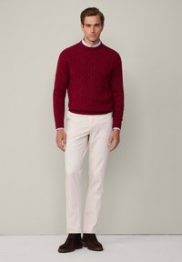 Roter Strickpullover mit Zopfmuster, cremefarbene, schmal geschnittene Hose und dunkelbraune Wildleder-Ankle-Boots, getragen von einem männlichen Model, das vor einem neutralen Hintergrund steht.