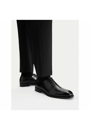 Schwarze, hochglanzpolierte Oxford-Lederschuhe, getragen mit schwarzer Anzughose und schwarzen Socken, seitlich und leicht von oben dargestellt.