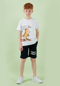 Garçon roux portant des baskets blanches, un short noir avec le texte "Protecteur de la planète" et un T-shirt blanc avec un dinosaure tenant une plante.