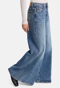 Korkean vyötärön leveälahkeiset siniset farkut. Ominaisuuksiin kuuluvat viisi taskua, nahkainen etiketti takana ja hapsuleveys. Tekstuurin näyttäisi olevan kestävä.