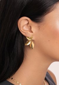 Femme aux cheveux lisses et foncés portant des boucles d'oreilles brillantes en forme de feuille d'or et un collier assorti en chaîne d'or.