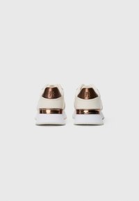 Une paire de baskets blanches avec des accents de talon bronze métallisé, vue de dos sur un fond blanc uni.