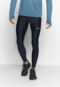 Granatowe legginsy sportowe wykonane z gładkiego, elastycznego materiału, z małym białym logo Nike na lewej udzie. Uzupełnione czarnymi butami do biegania.