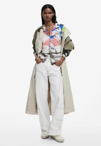 Trenchcoat in kaki, gelaagd over een kleurrijk gedrukt shirt en witte wijdvallende jeans met zichtbare stiksels. Schoeisel is licht van kleur en textuur.