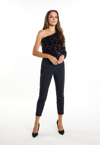 Top nero a spalla unica con paillettes, abbinato a pantaloni neri aderenti e scarpe con tacco alto nere, che mostrano una texture liscia e un design aderente.