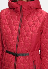 Rød kviltet jakke med hætte, prydet med et geometrisk mønster, en lynlås foran og et sort bælte. Glat tekstur og isoleret design.