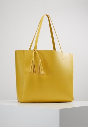 Borsa tote gialla in pelle con due manici lunghi, una texture liscia e frange decorative su un lato, che presenta un design semplice e minimalista.