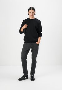 Svart bomullströja med ribbad mudd vid ärmar och nederkant, kombinerad med grå denimjeans. Modellen bär svarta sneakers med vita detaljer.