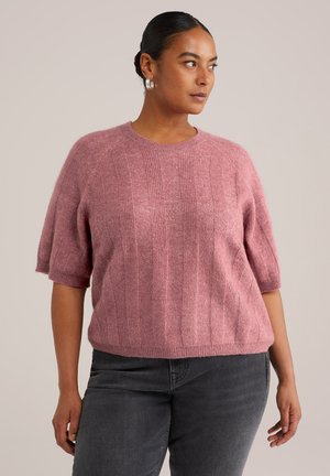 Rosa Strickpullover mit kurzen Ärmeln, der über eine vertikale Rippstruktur und einen runden Halsausschnitt verfügt. Kombiniert mit dunkelgrauen Jeans.