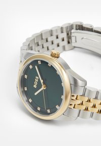 Relógio de pulso com pulseira de metal dourado e prateado, mostrador verde escuro, ponteiros dourados, marcadores de horas em cristal e luneta dourada, indicando as 8:55:30.