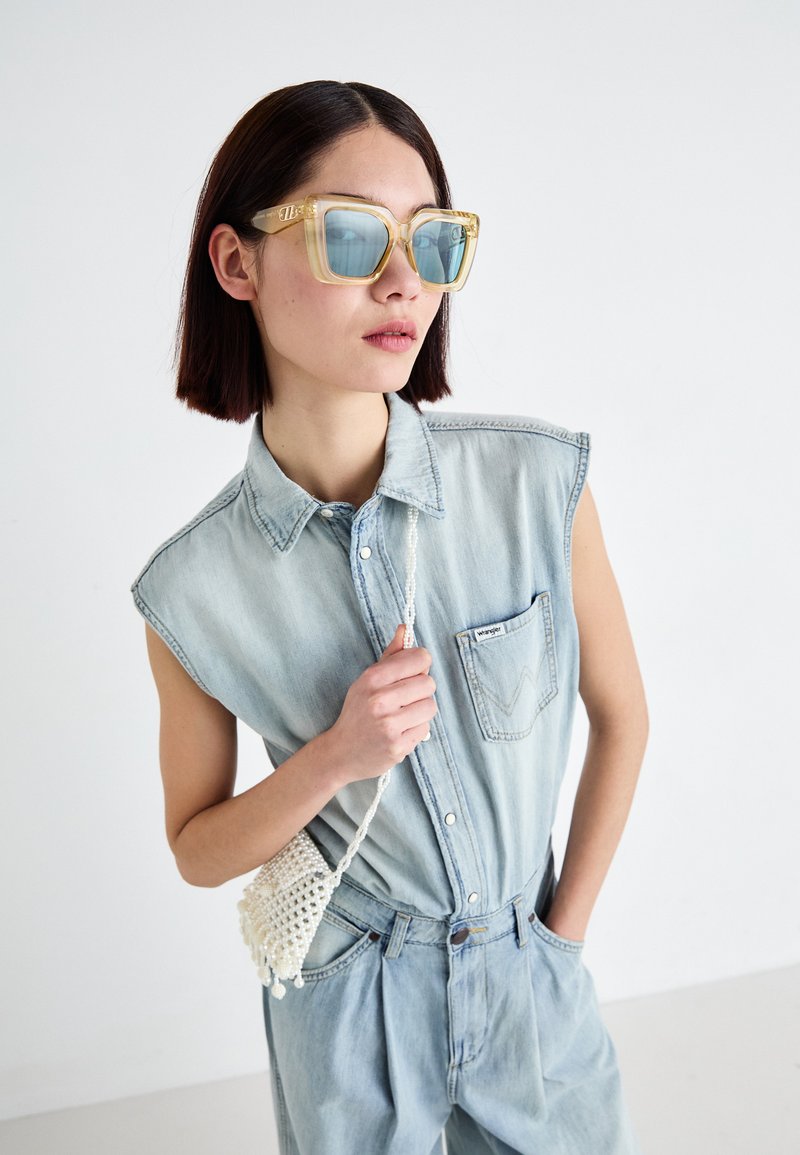 Jeans-Overall in Hellblau mit Kragen und kurzen Ärmeln, ausgestattet mit einer Brusttasche. Das Modell hält eine weiße Häkelhandtasche.