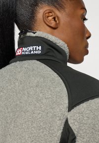 Chaqueta de forro polar con un cuerpo gris y detalles en negro, cuenta con un alto cuello y un parche de logo en la parte posterior, tejido texturizado y un diseño elegante.