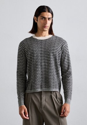 Ein schwarz-weißer Strickpullover mit wiederholtem Textmuster, Rundhalsausschnitt und gerippten Bündchen. Kombiniert mit beigen, plissierten Hosen.