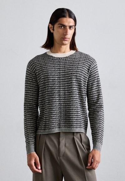 Un pull en tricot noir et blanc présente un motif de texte répété, un col en rond et des poignets côtelés. Associé à un pantalon plissé beige.