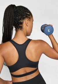 Črna športna modrc z nizkim hrbtom, z tankimi trakovi in gladko teksturo. Oseba drži modro utež, označeno s številko 3.