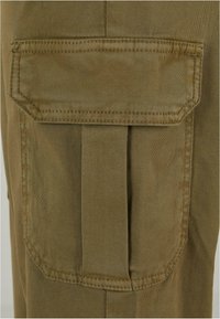 Gros plan d'une poche cargo en tissu kaki avec rabat et détail plissé, cousue sur un tissu assorti.