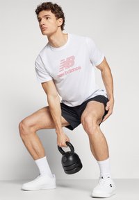Balta T-kreklu ar sarkano "New Balance" logotipu, melni šorti, baltas zeķes un baltas kurpes. Individuāls tur melnu kettlebellu un izpilda pietupienu.