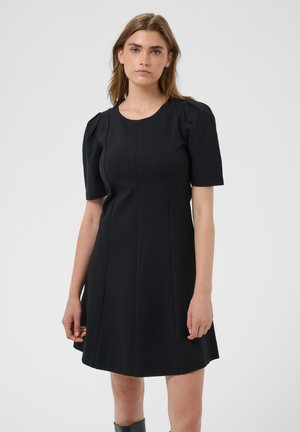 Femme portant une robe noire courte à manches courtes, longueur genoux, avec un détail froncé à l'épaule, debout devant un fond gris clair uni.