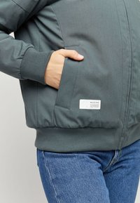 Graue Bomberjacke mit strukturierter Oberfläche, gerippten Bündchen und Bund. Verfügt über eine Seitentasche und einen weißen Logo-Patch nahe dem Saum.