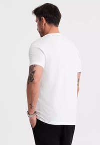 Hvid bomulds t-shirt med korte ærmer, rund hals og en simpel design, båret af en mand med synlige tatoveringer på armen.