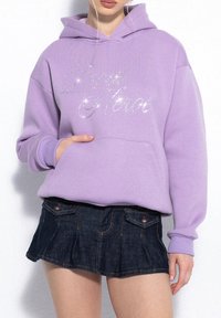 Fioletowa bluza z kapturem z błyszczącą srebrną grafiką „Dobry Bohater” z przodu. Posiada kieszeń z przodu i luźny krój, zestawiona z ciemnoniebieską mini spódnicą.